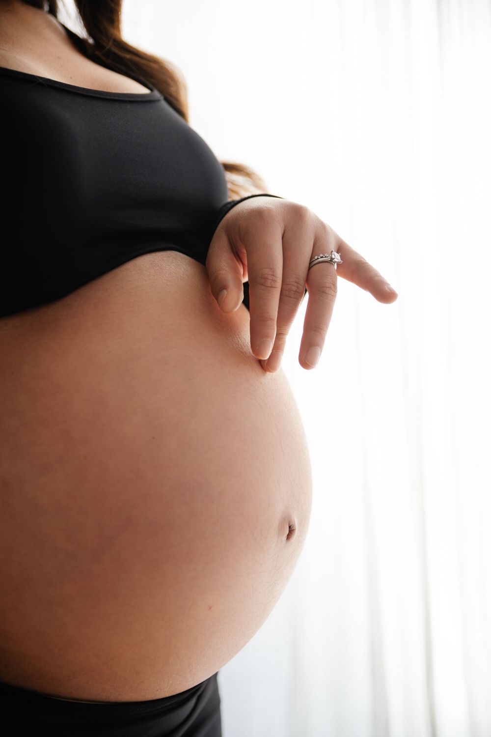 Pregnant woman in black crop top gently touching her bare baby bump, standing near a bright window.
