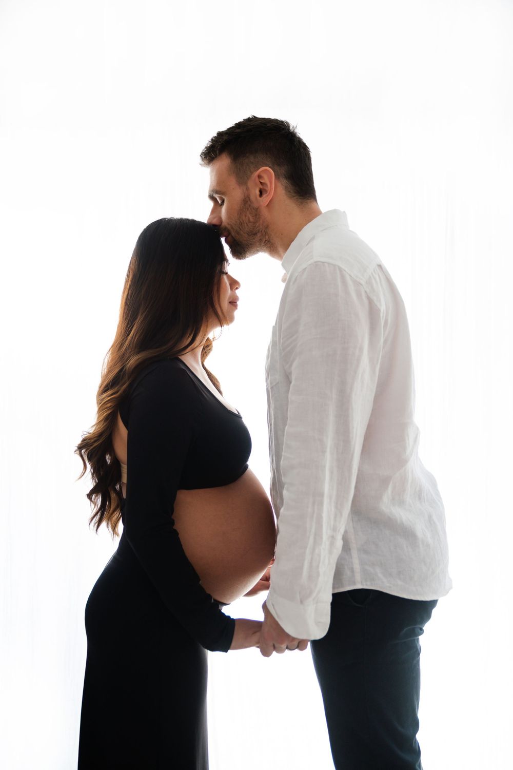 Silhouette of man kissing pregnant woman's forehead while holding hands against white background.