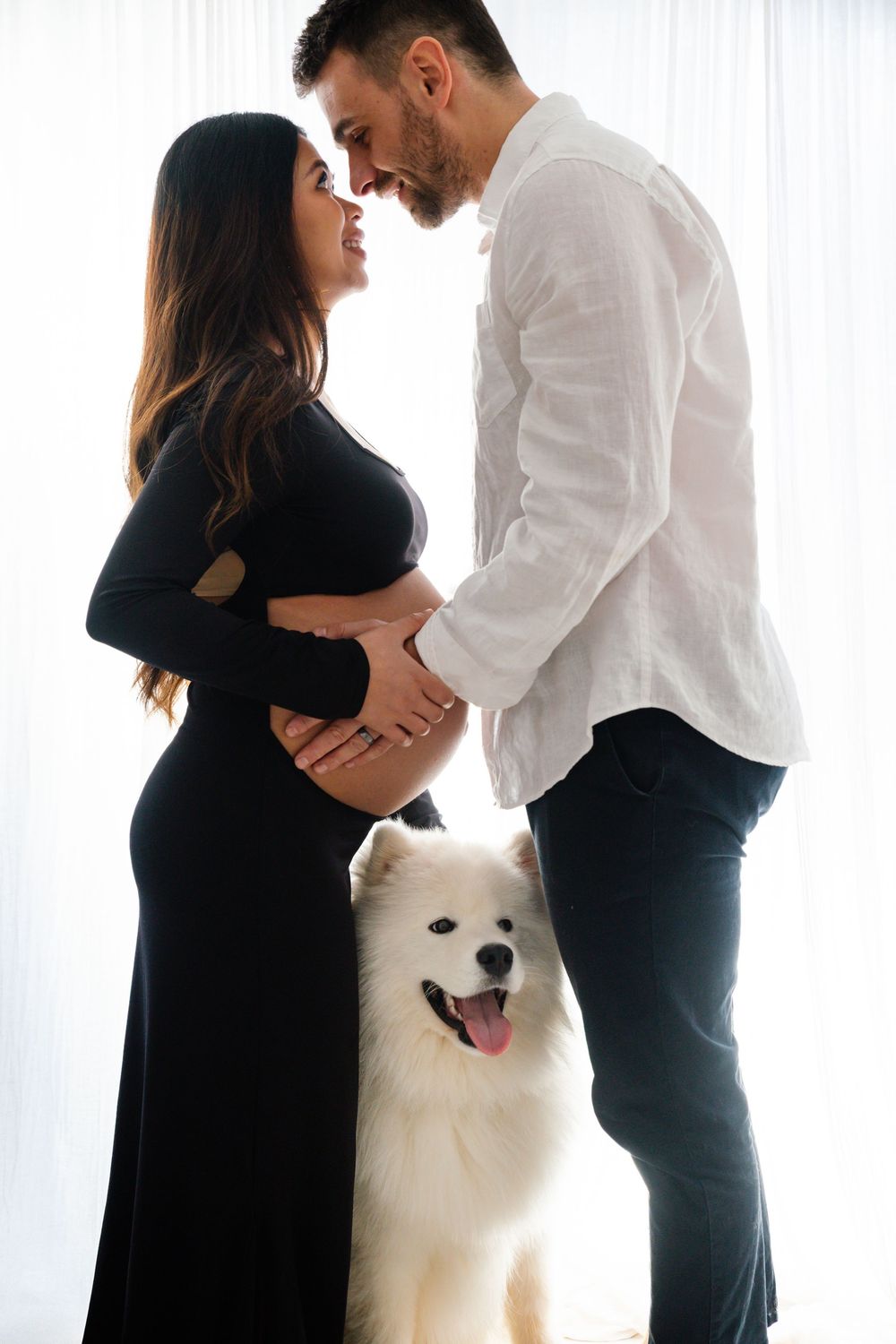 Pregnant couple in black and white outfits sharing a tender moment with a fluffy white Samoyed dog.