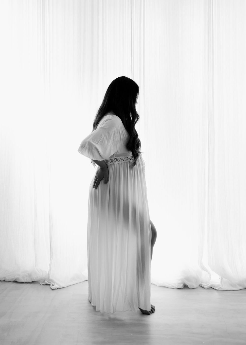 Pregnant woman in white sheer maternity gown standing sideways in soft light, black and white portrait photography.