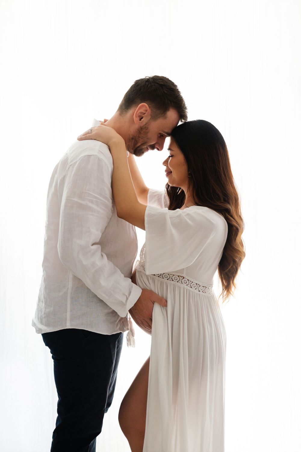 Couple in white outfits share an intimate forehead touch during a maternity photoshoot.