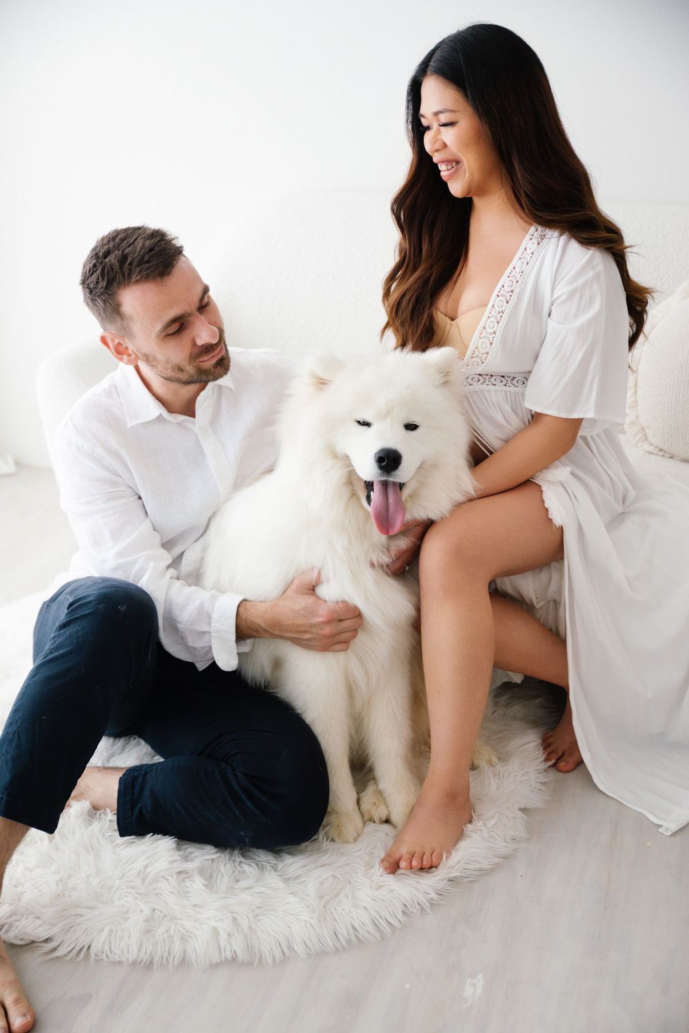 Happy couple in white outfits petting a fluffy white Samoyed dog on a fur rug in a bright room.