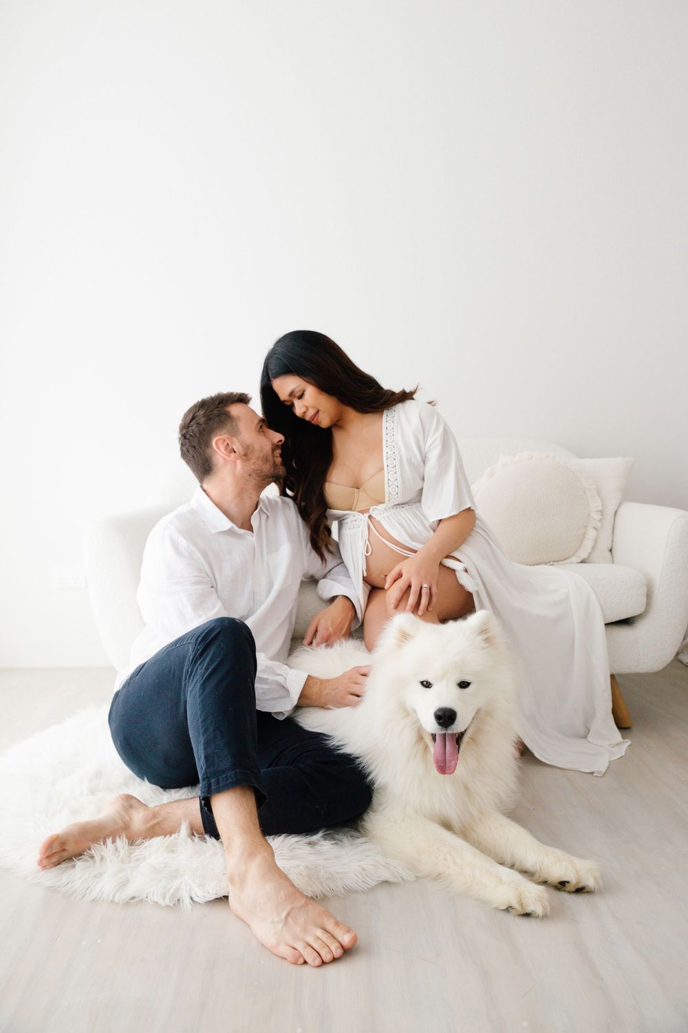 Couple sitting on floor with fluffy white Samoyed dog in bright white room, woman in white dress on couch.
