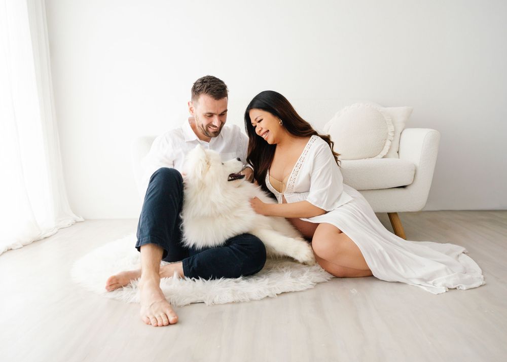 Couple sitting on white fur rug with large fluffy white dog in a bright minimalist studio.