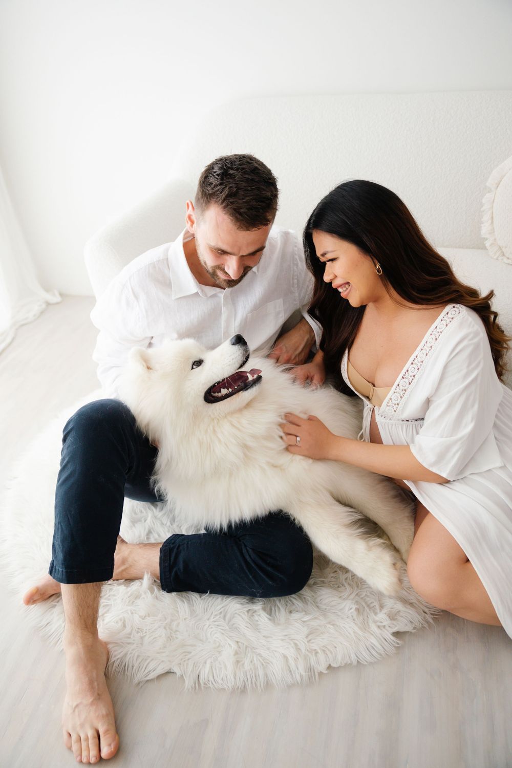 Happy couple sitting on fur rug petting their white Samoyed dog in a bright white room.