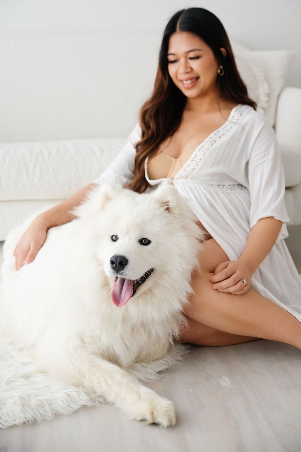 Pregnant woman in white robe sitting on floor smiling while petting a fluffy white Samoyed dog indoors.