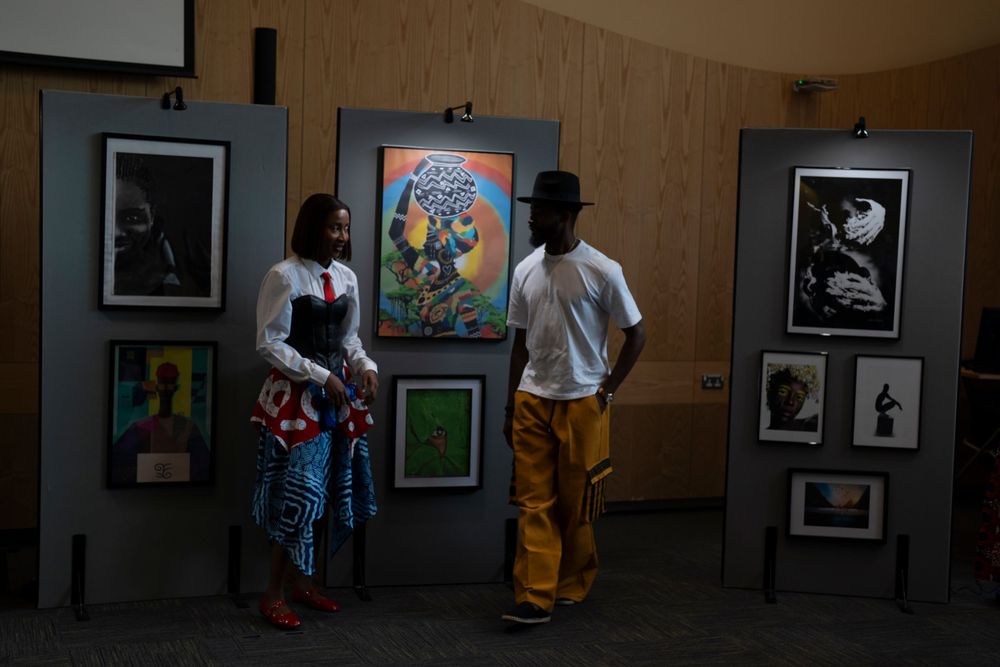 Two people converse at an art gallery exhibition featuring colorful paintings and black-and-white photographs on display panels.