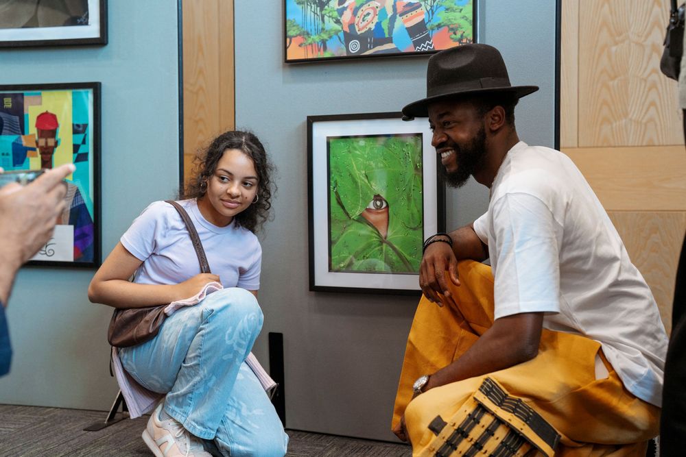 Two people crouch and chat at an art gallery, surrounded by colorful paintings on display.