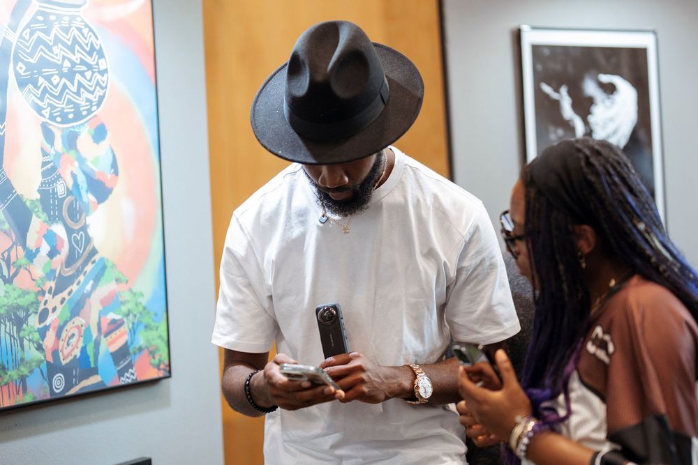 Two people in an art gallery look at a phone, one wearing a black wide-brim hat and white shirt.
