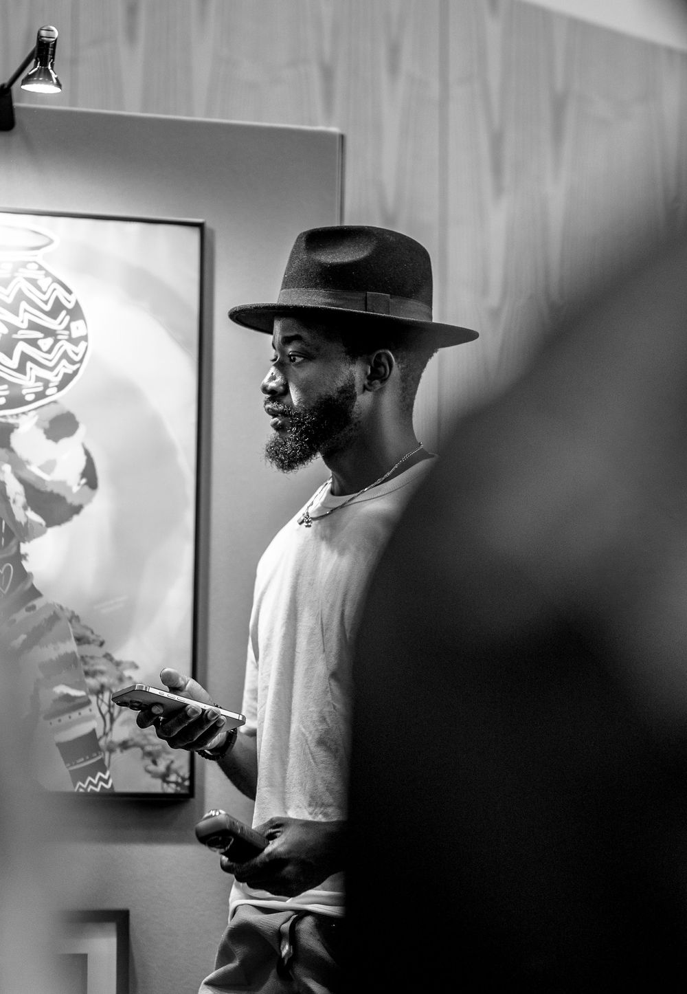 Black and white photo of a bearded man wearing a wide-brim hat holding a phone at an art gallery event.
