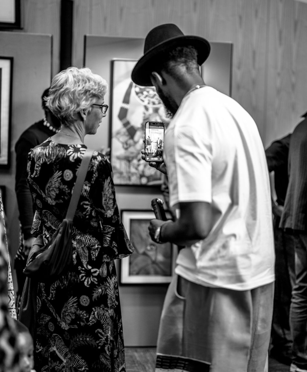 Black and white photo of two people viewing art at a gallery, one wearing a hat and holding a smartphone.