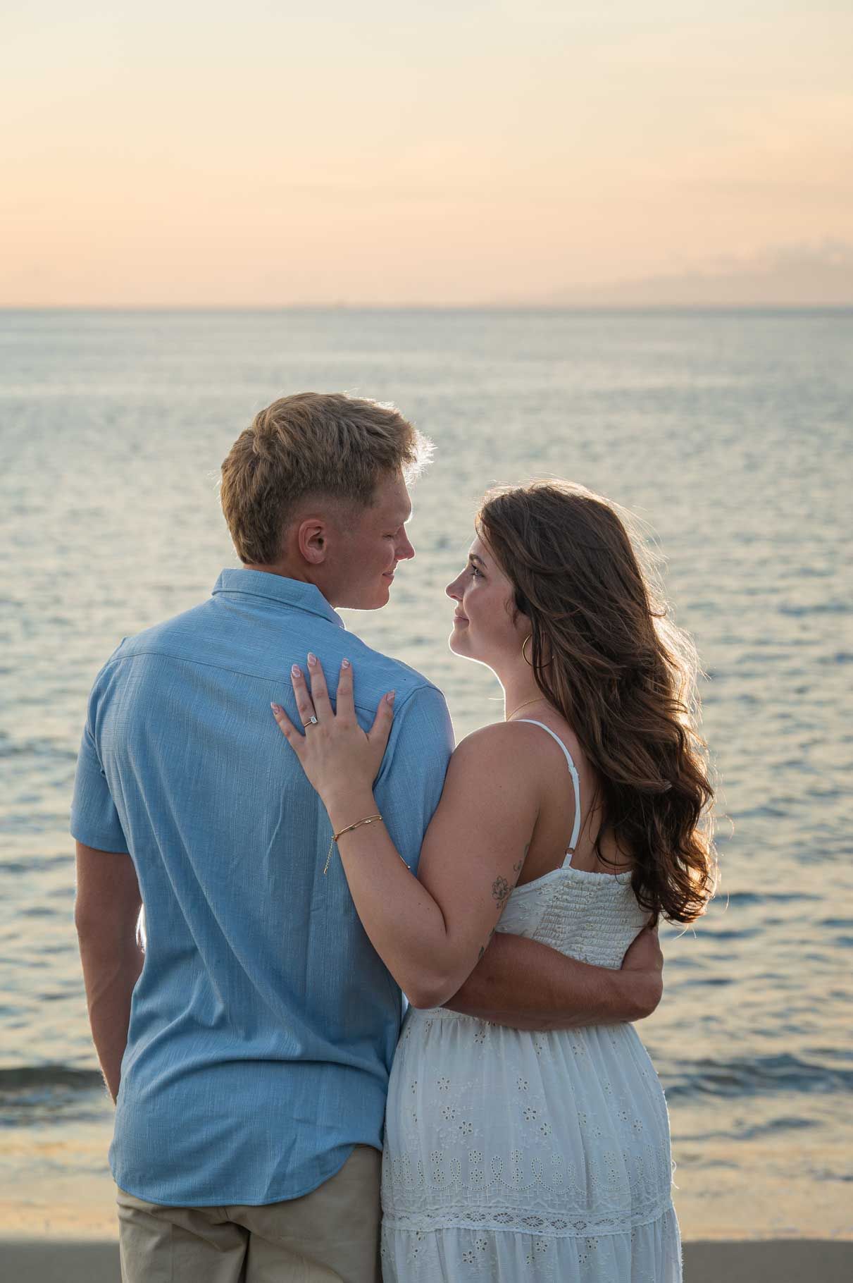 Couple embracing at sunset by the ocean, woman showing engagement ring, wearing white dress and blue shirt.