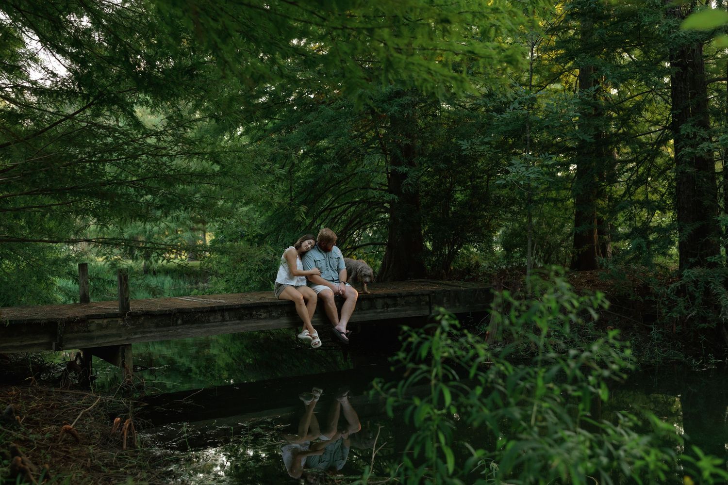 Two people sit together on a wooden bridge surrounded by lush green trees, reflected in the calm water below.