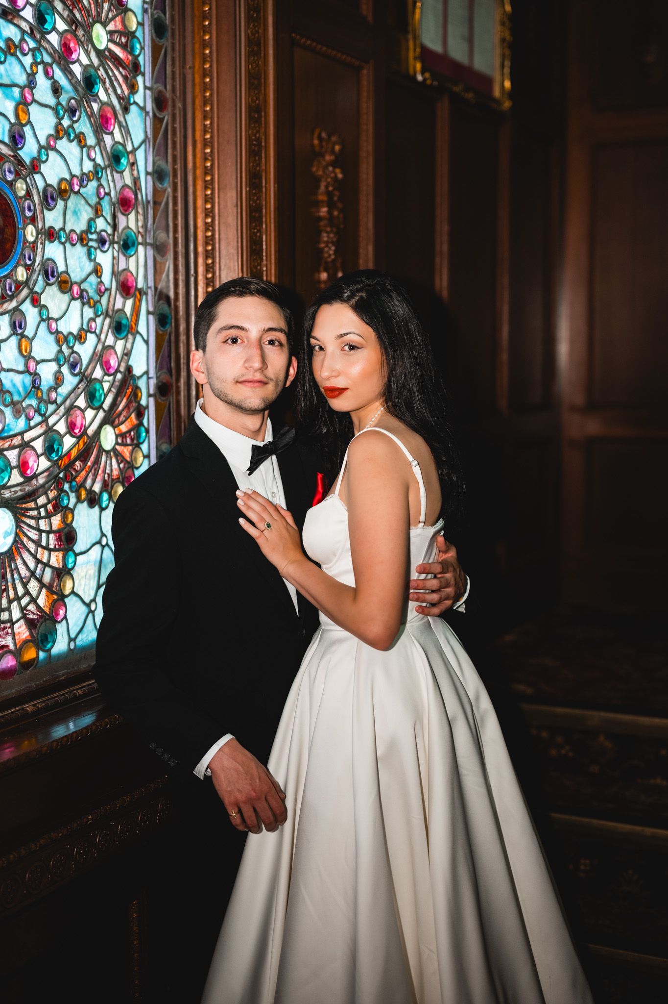 Elegant bride and groom posing together in front of a colorful stained glass window in a grand venue.