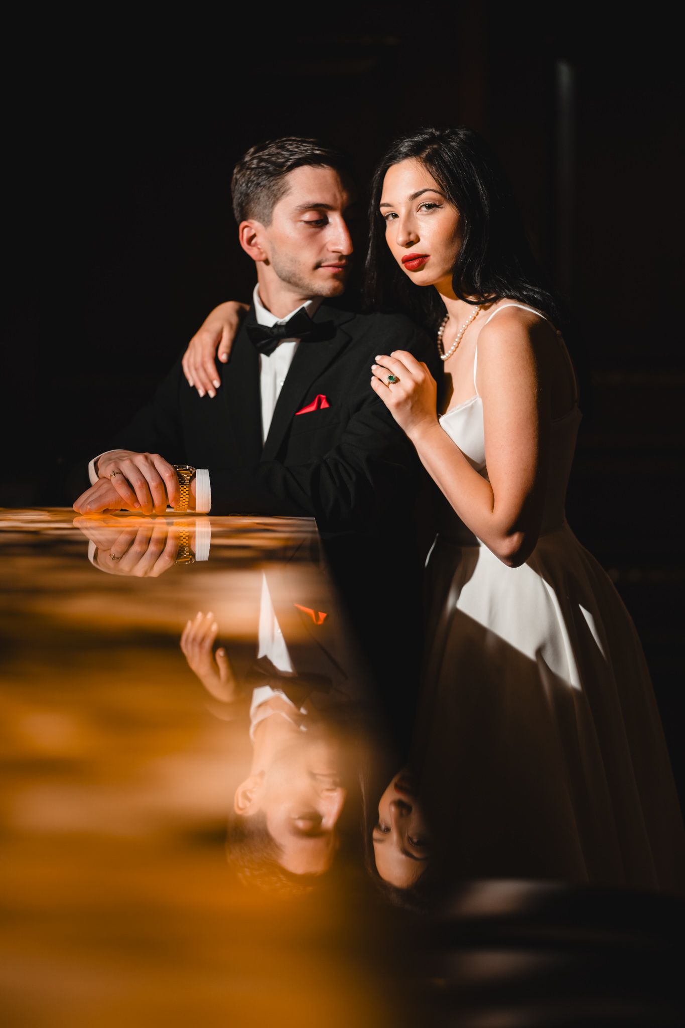 Elegant couple in formal attire pose at a bar, man in tuxedo and woman in white gown with red lips.