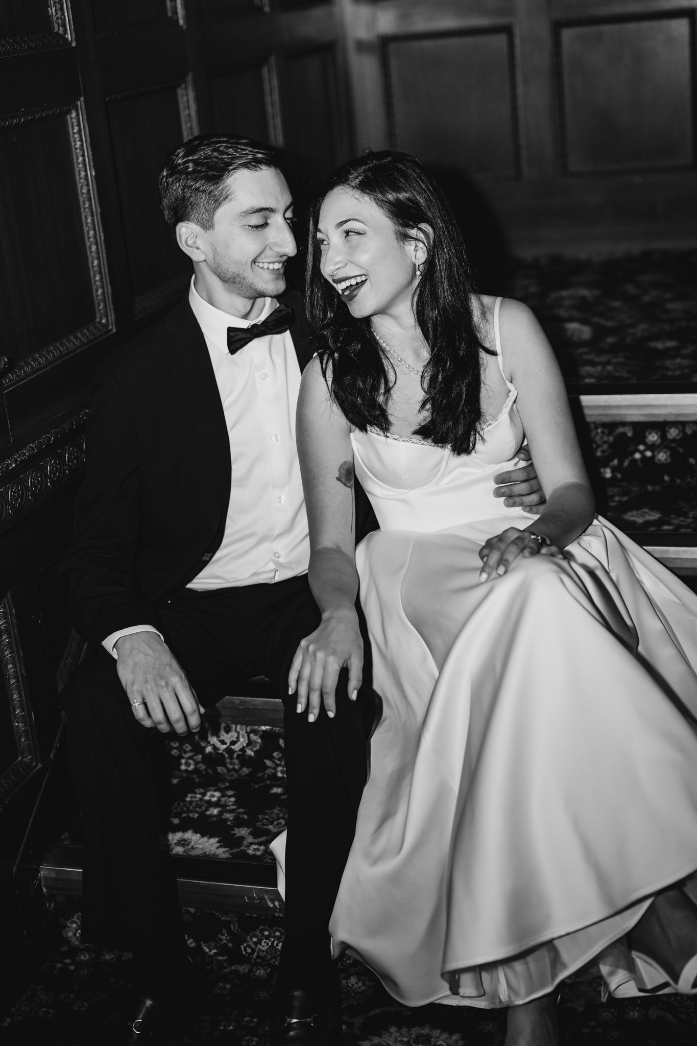 Black and white photo of a laughing bride in white dress and groom in tuxedo sitting together on a bench.
