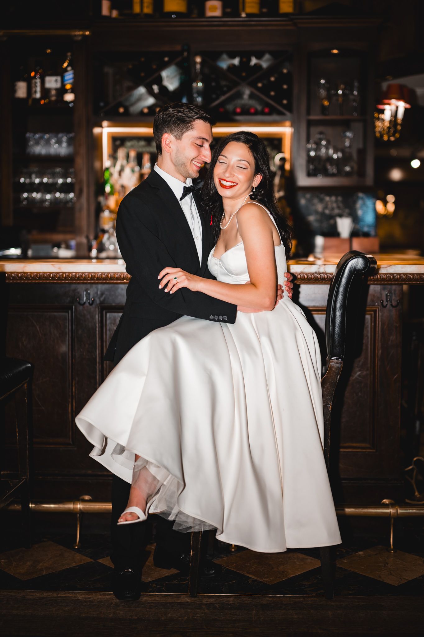Bride and groom embrace at a dark wood bar, she in a white tea-length gown, he in a black tuxedo.