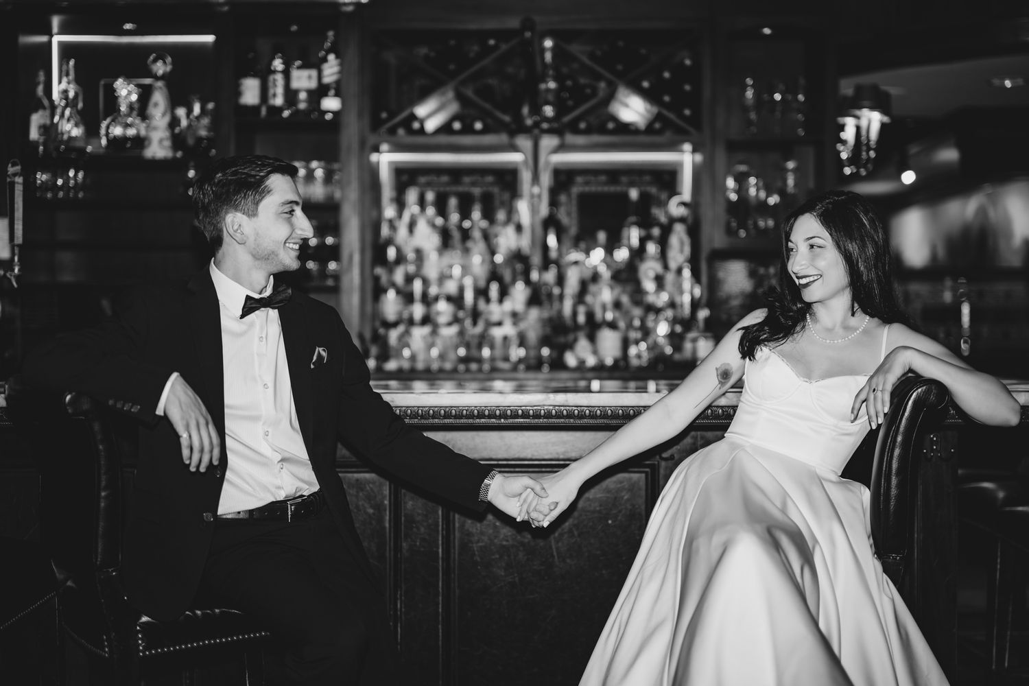 Black and white photo of a couple in wedding attire holding hands and smiling at each other in an elegant bar setting.