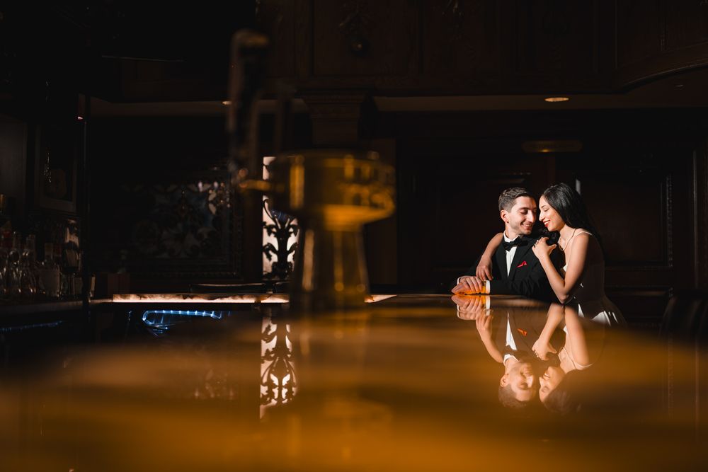 Couple sharing romantic moment at dimly lit bar, reflected in glossy surface, dressed in elegant formal attire.