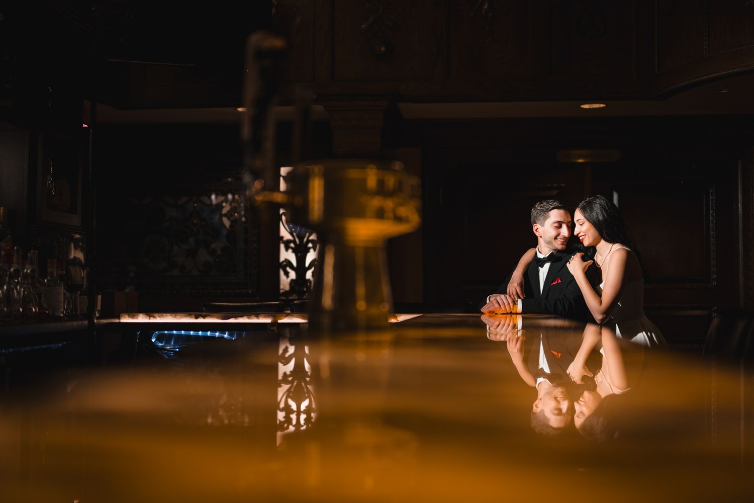 Couple sharing romantic moment at dimly lit bar, reflected in glossy surface, dressed in elegant formal attire.
