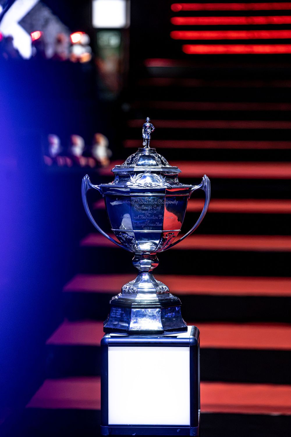 Gleaming silver trophy cup with lid displayed on white pedestal under dramatic blue and red stage lighting.