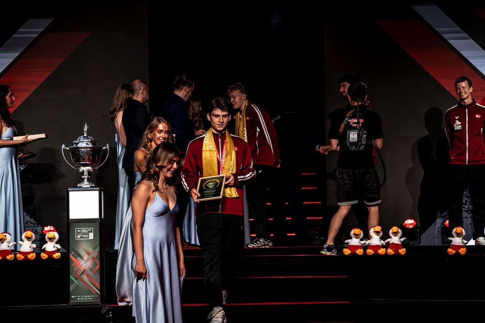 Young man in gold scarf receives award on stage at formal ceremony with trophy and elegantly dressed attendees.