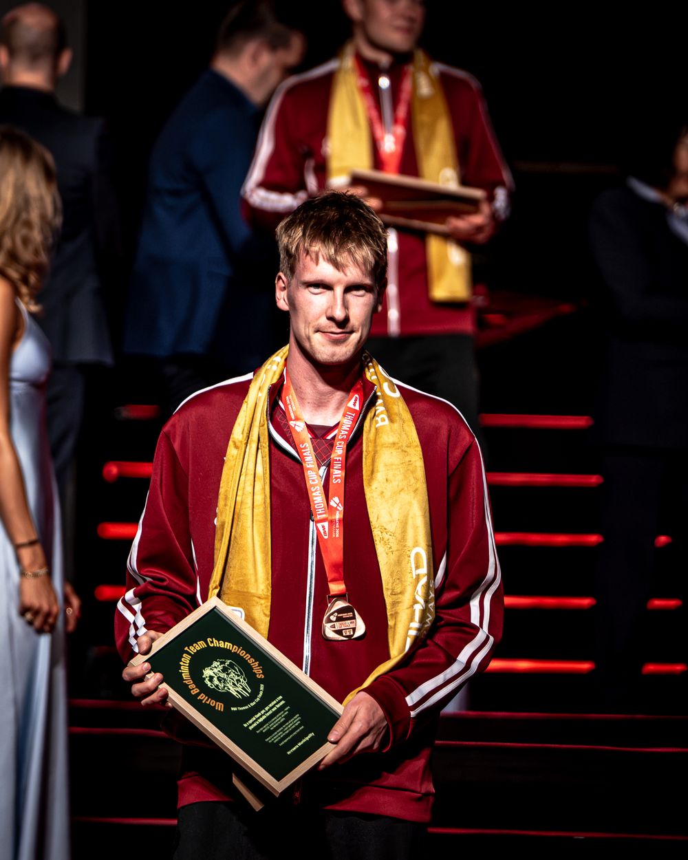 Young male athlete in red jacket holds award plaque, wearing gold scarf and medal at a sports ceremony.