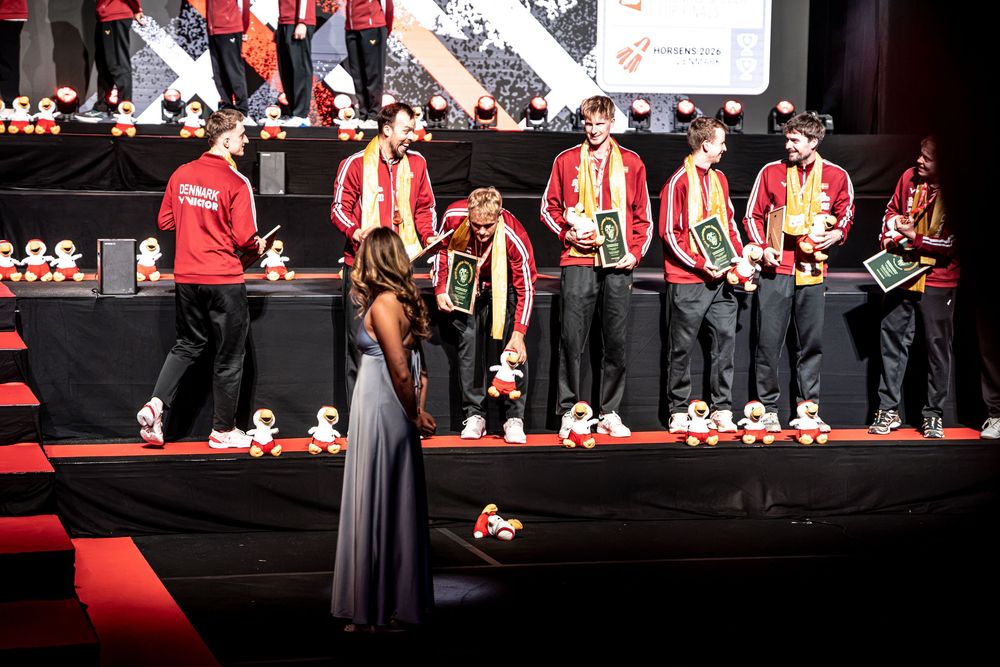 Award ceremony on stage with athletes in red uniforms receiving trophies from a presenter in a gray gown.