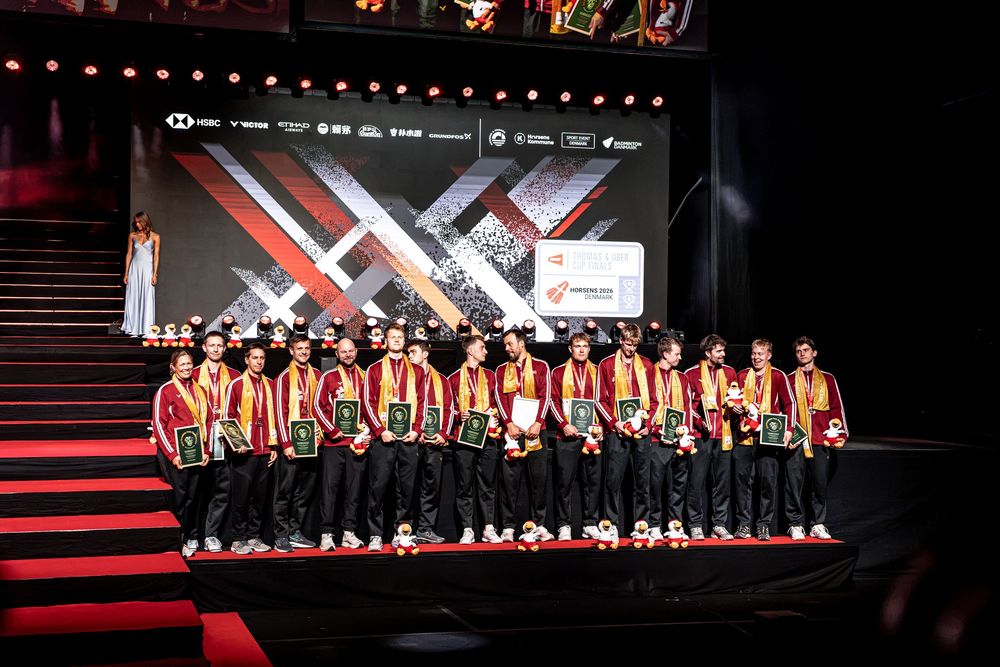 Team in red and gold uniforms poses on stage at a badminton championship awards ceremony.