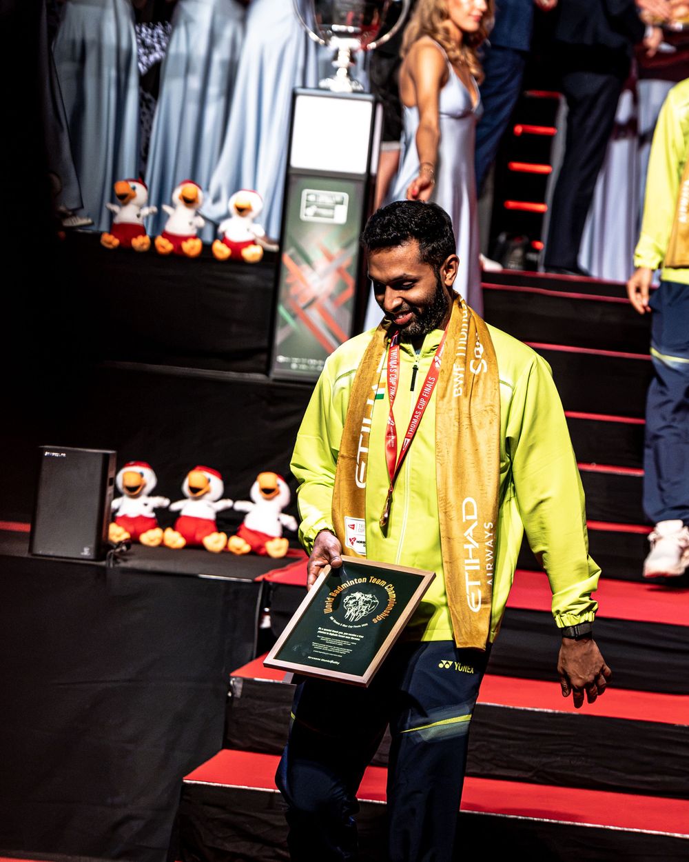 Man in yellow Yonex jacket holding award plaque at badminton tournament ceremony with trophies in background.