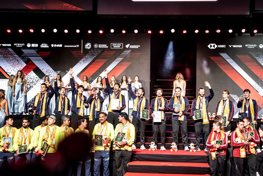 Athletes celebrate on stage at a badminton championship awards ceremony, wearing gold sashes under colorful stage lighting.