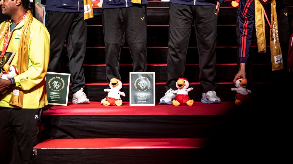 Badminton tournament podium with stuffed animal mascots, trophies, and plaques displayed on red stairs.