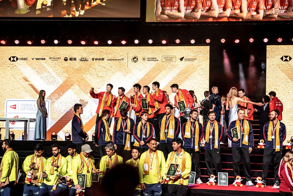 Athletes in red and yellow uniforms celebrate on a decorated stage at an international badminton championship awards ceremony.