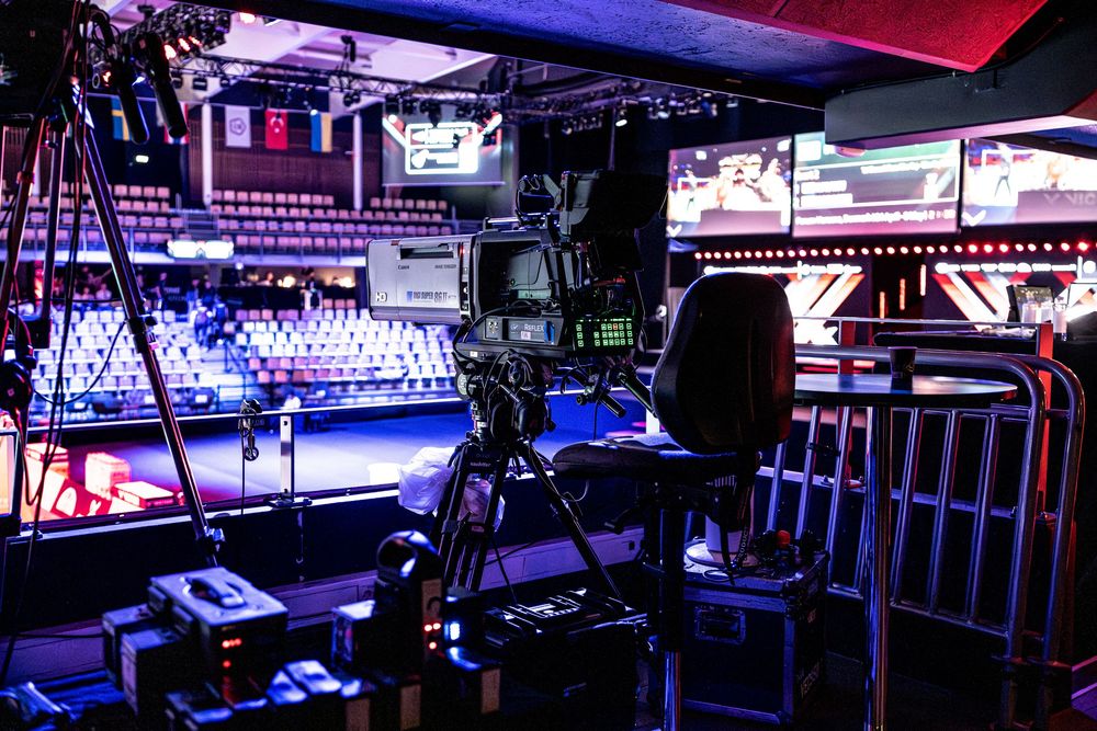 Professional TV broadcast camera setup on tripod in arena venue with monitors and production equipment under purple lighting.