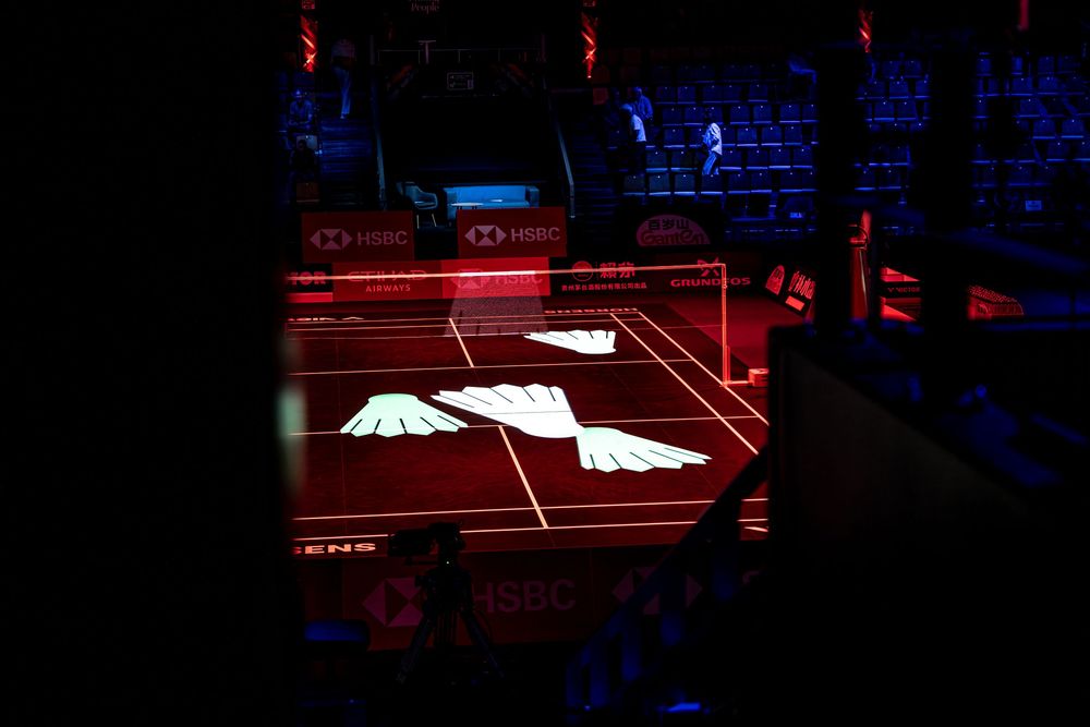 Badminton court illuminated with glowing wing logo projection during HSBC tournament opening ceremony in dark arena.