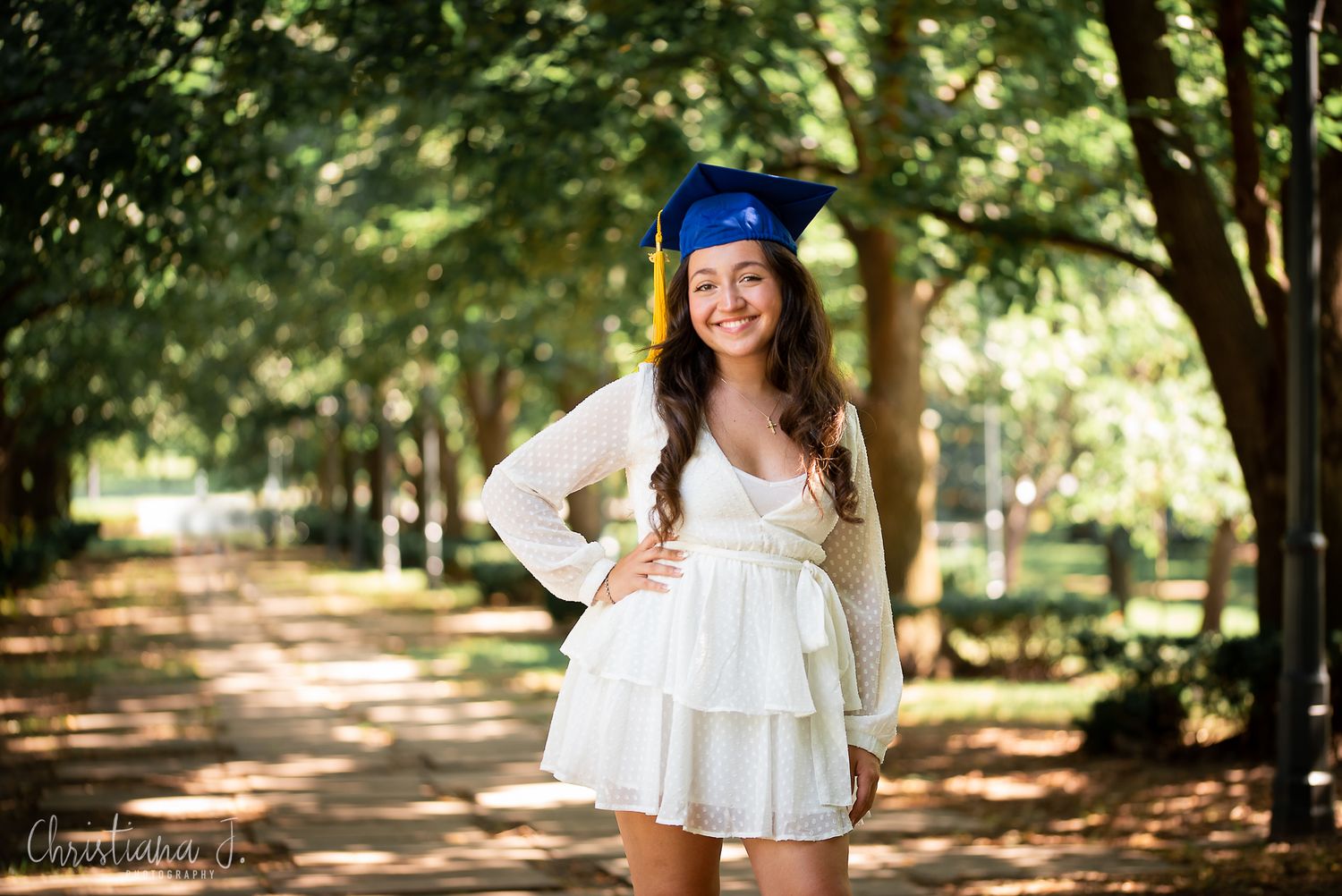 Smiling graduate in white dress and blue cap standing on a tree-lined pathway.