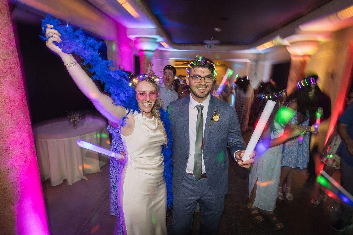 Couple celebrating at wedding reception with glow sticks and colorful lighting, smiling and dancing with guests in background.