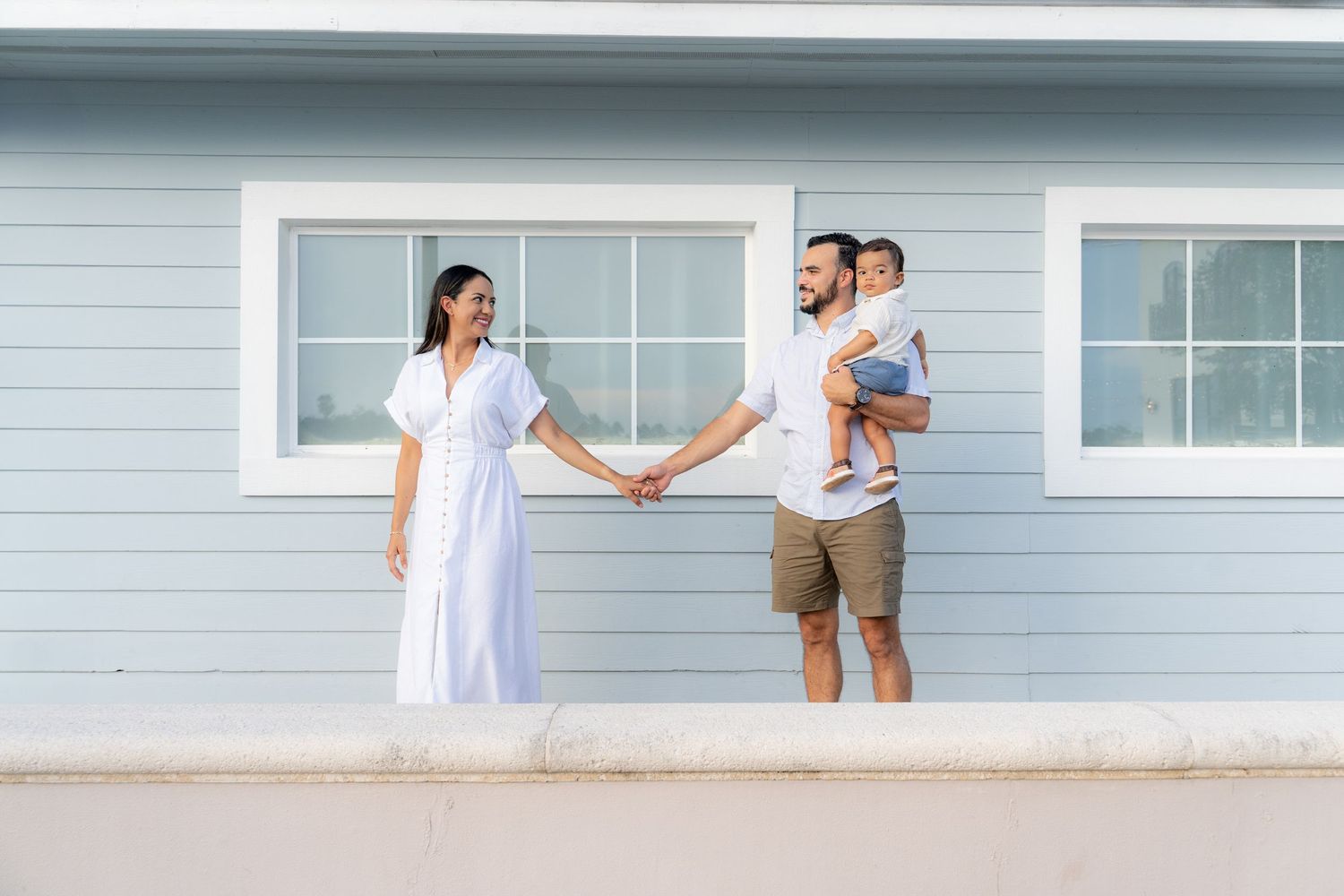 A young family holding hands outside a light blue beach house, with father carrying a toddler.