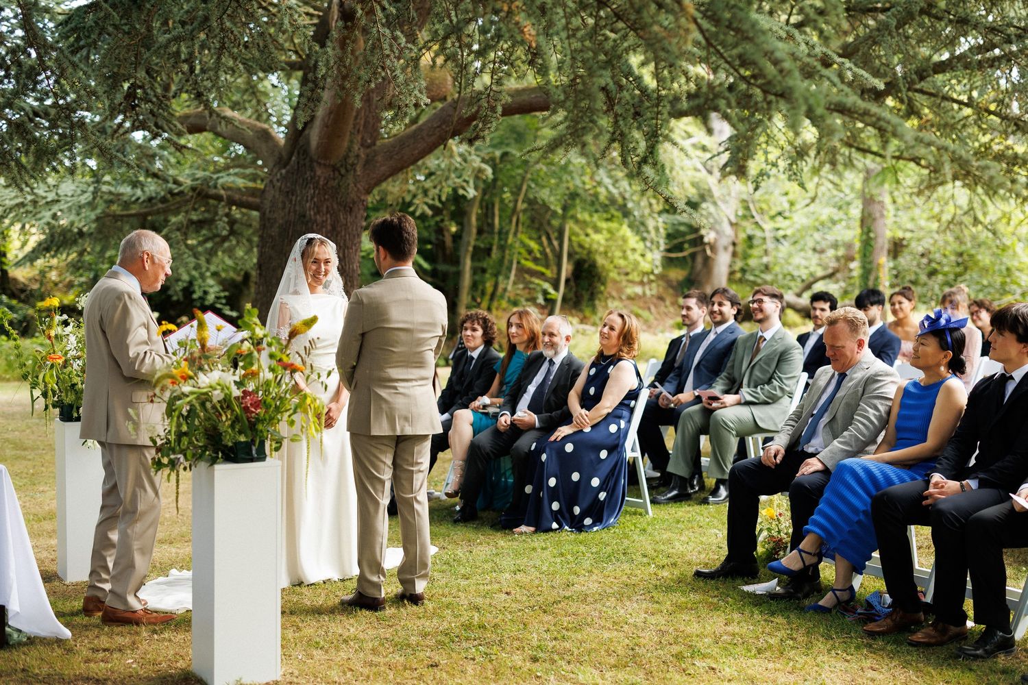 Outdoor wedding ceremony under a large tree with bride and groom at the altar, officiant reading, and seated guests on a sunny day.