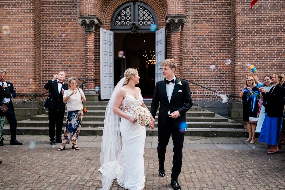 Bride and groom exit brick church amid bubbles, guests celebrating their wedding ceremony outdoors.