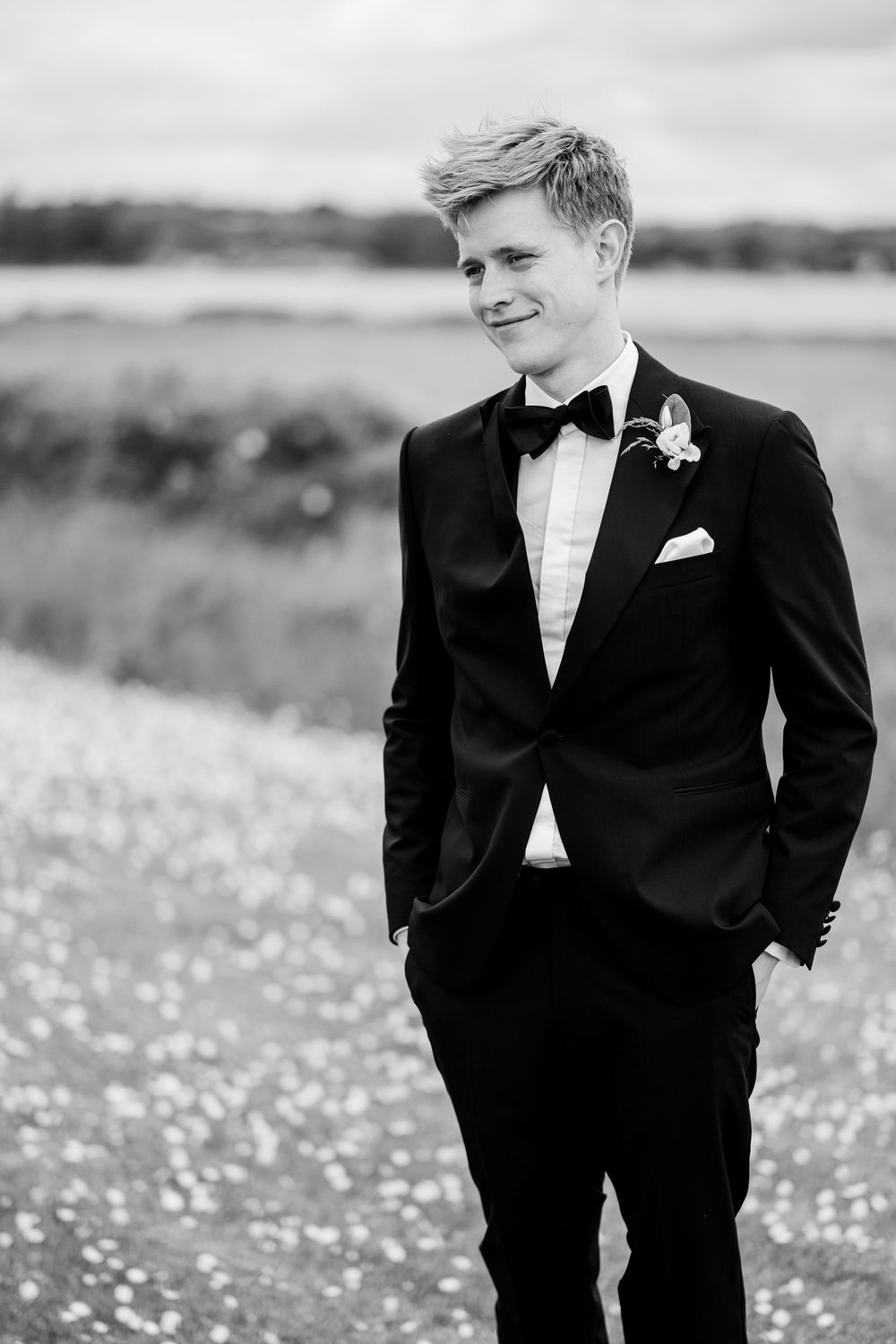 Young groom in black tuxedo with bow tie and boutonniere stands in field, black and white wedding portrait.
