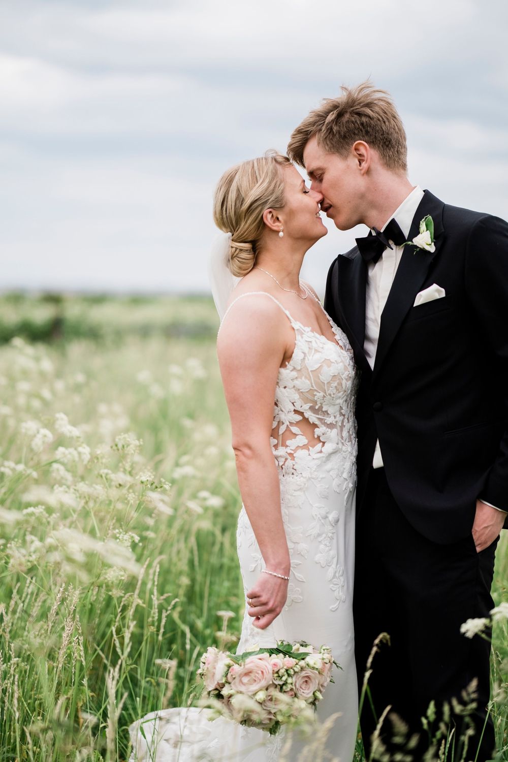 Bride and groom share a romantic almost-kiss in a lush green field, bride holding a soft pink bouquet.