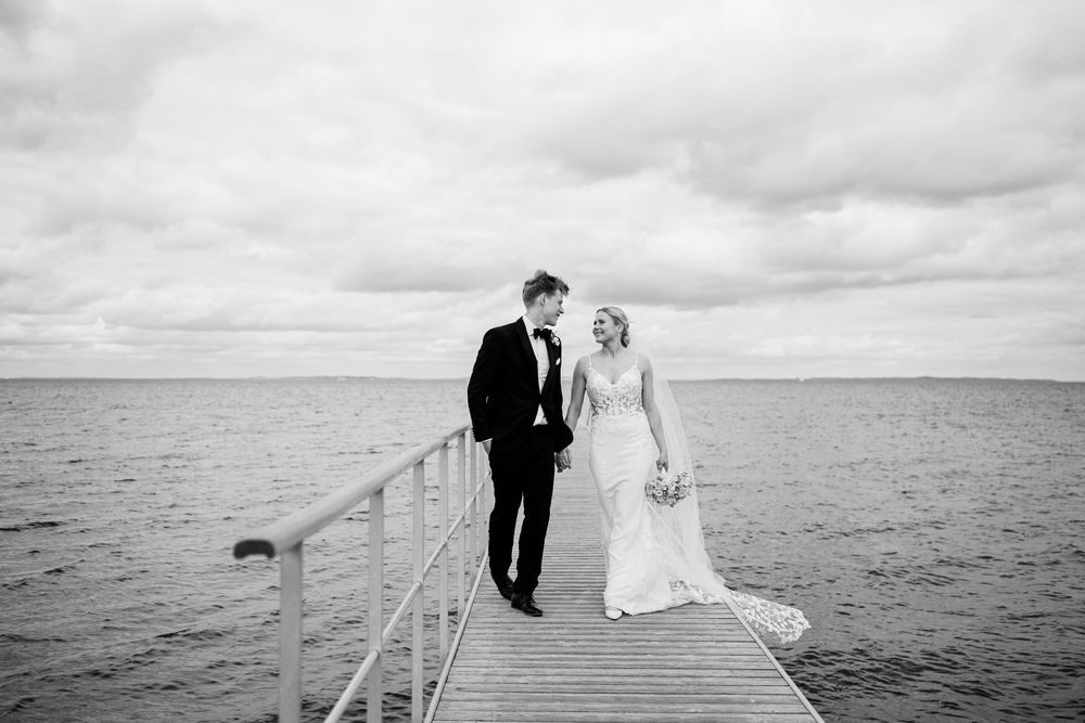 Bride and groom walk hand in hand on pier over calm ocean waters in black and white wedding photo.