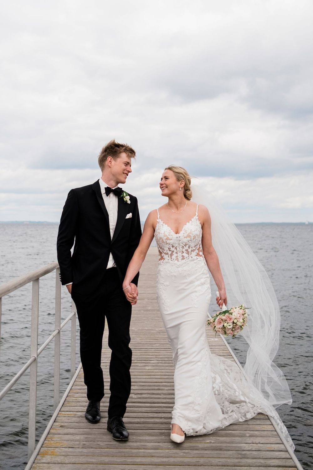 Bride and groom walk hand-in-hand on a pier by the sea, she in a lace gown, he in a black tuxedo.