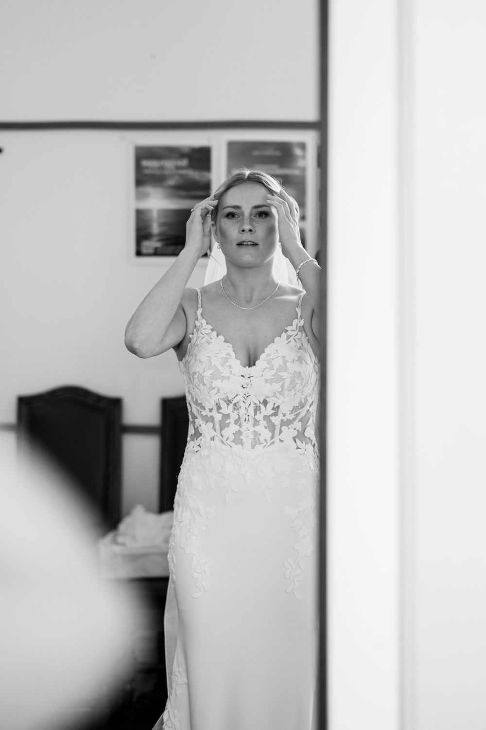 Black and white photo of a bride in a lace wedding gown adjusting her hair while gazing into a mirror.