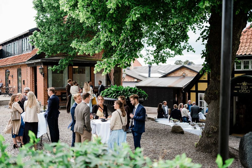 Guests mingle outdoors at an elegant garden venue with rustic buildings and lush green trees during a summer event.