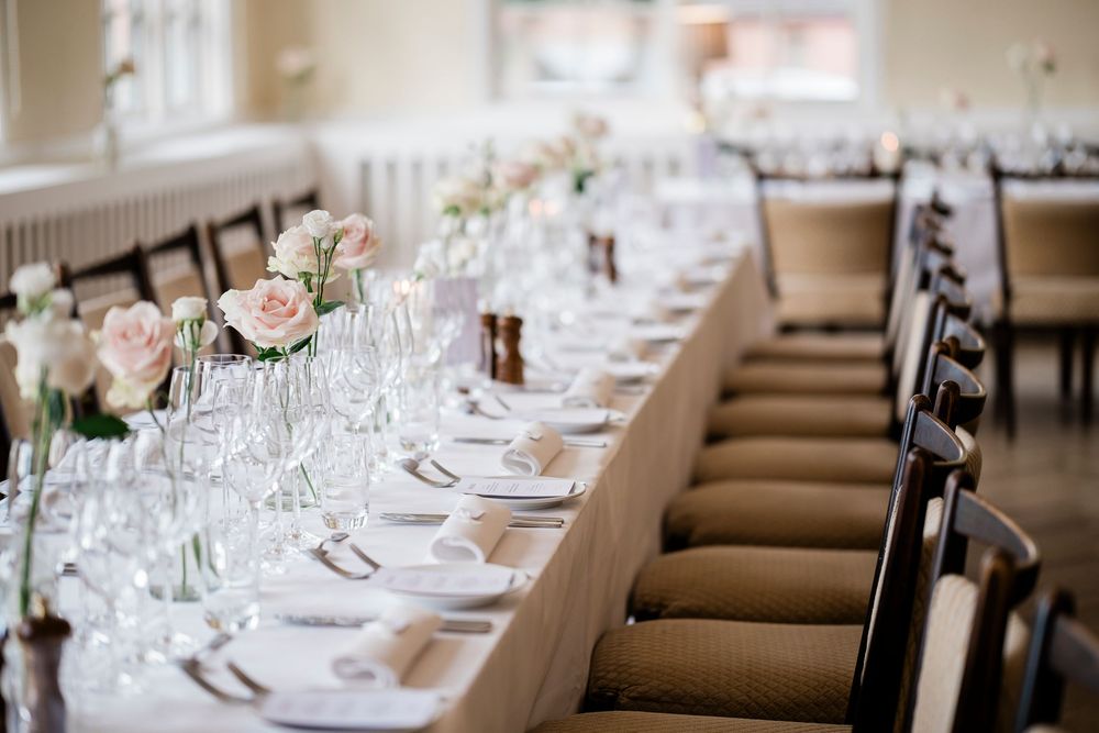 Elegant wedding reception table with white linen, pink roses, crystal glasses, and dark wood chairs in a bright venue.