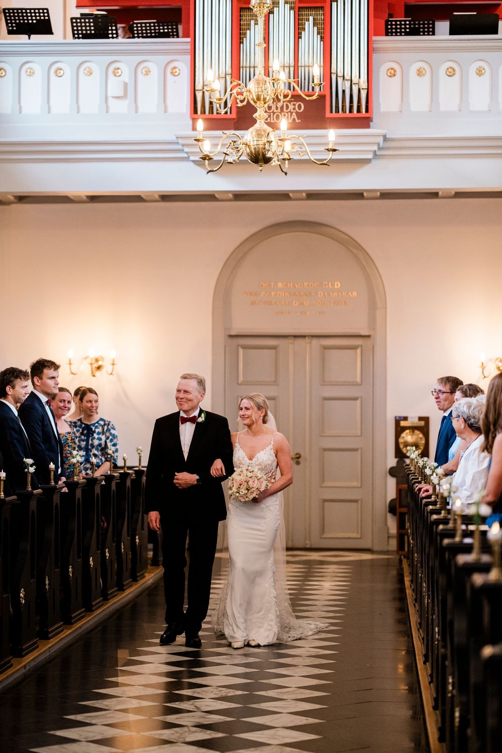 Bride walks down church aisle with father, wearing lace gown, holding bouquet, guests seated in pews, pipe organ above.
