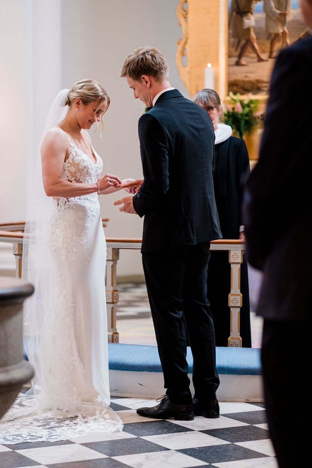 Bride places wedding ring on groom's finger during church ceremony with officiant in background.