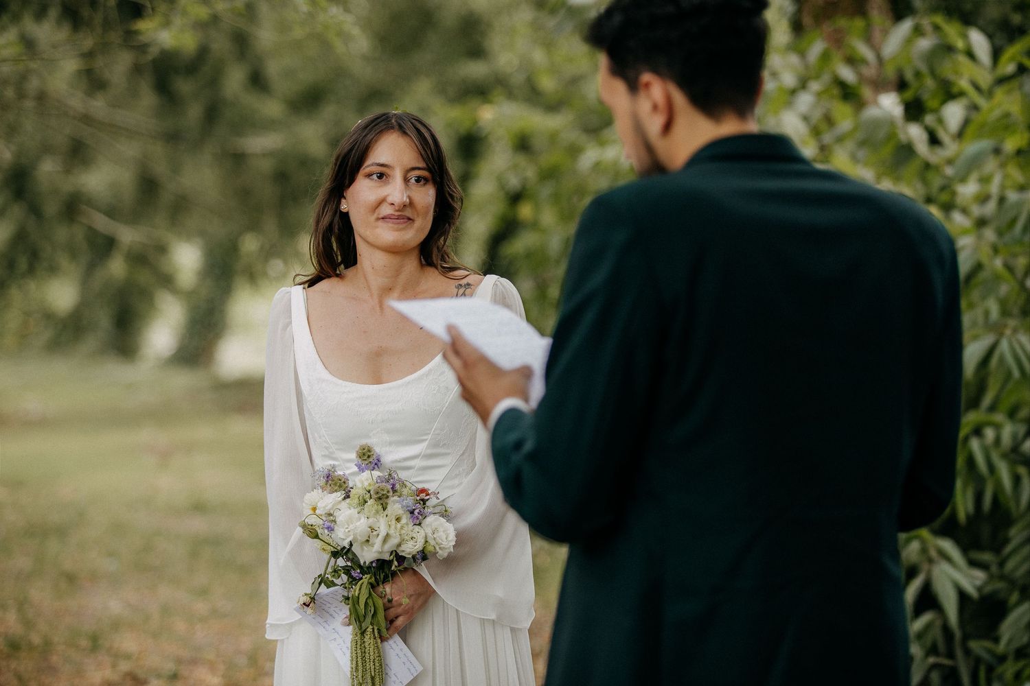 Bride in white dress holds floral bouquet while groom reads wedding vows outdoors among green trees.